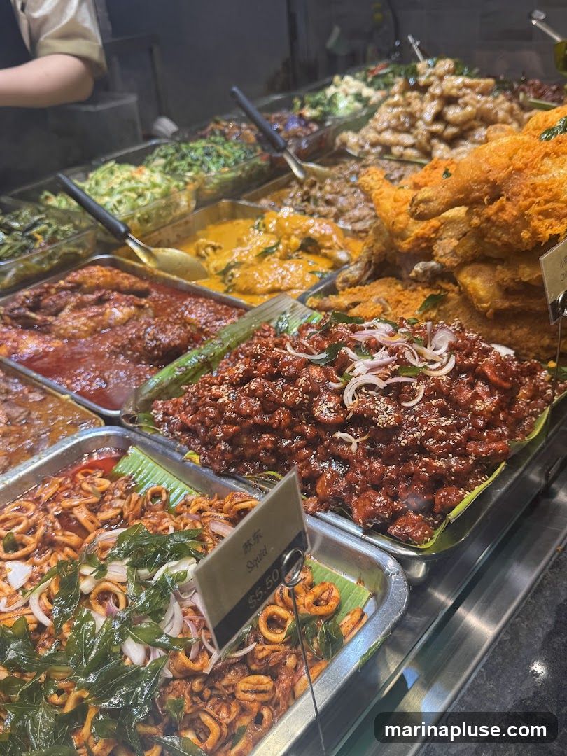 A close-up view of a food display case filled with various dishes. In the foreground, a tray of squid with onions and herbs is visible, with a sign indicating "Squid $5.50". Behind it, a tray of dark, glazed meat is topped with sesame seeds and onions. Further back, a variety of curries, stir-fried vegetables, and fried chicken are arranged in metal trays. A person's arm and shoulder are visible on the left, suggesting a food service counter.