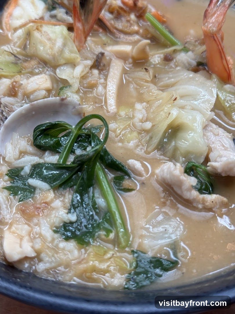 A close-up shot of a hearty stew filled with seafood and vegetables. Visible ingredients include shrimp, clams, leafy greens, cabbage, and what appears to be fish or chicken pieces, all in a creamy broth.