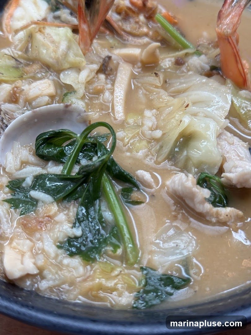 A close-up shot of a hearty stew filled with seafood and vegetables. Visible ingredients include shrimp, clams, leafy greens, cabbage, and what appears to be fish or chicken pieces, all in a creamy broth.
