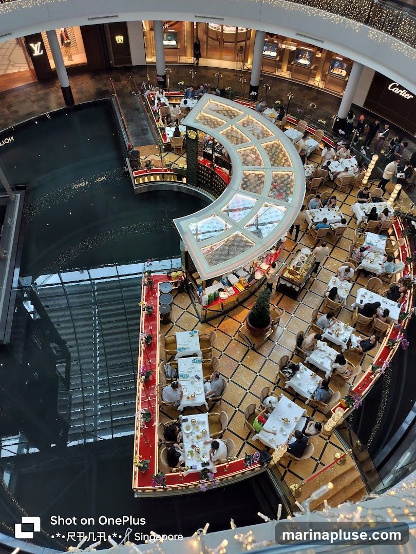 An aerial view of a bustling indoor dining area situated around a canal. Several tables are occupied by diners enjoying their meals. The dining area features elegant decor with red accents and floral arrangements. In the background, luxury brand stores like Louis Vuitton and Cartier are visible.