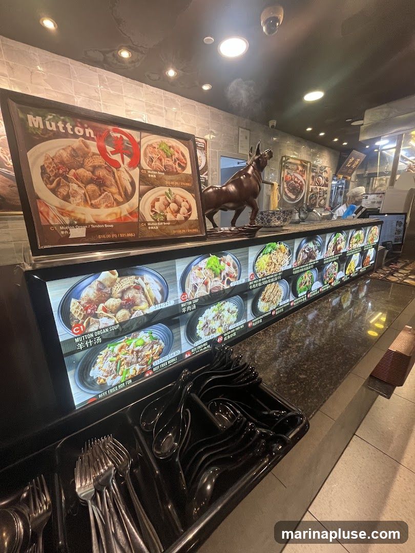 A display of food items with prices, featuring mutton dishes and noodle dishes. A statue of a bull stands prominently on the counter. Cutlery is arranged in a tray in the foreground.