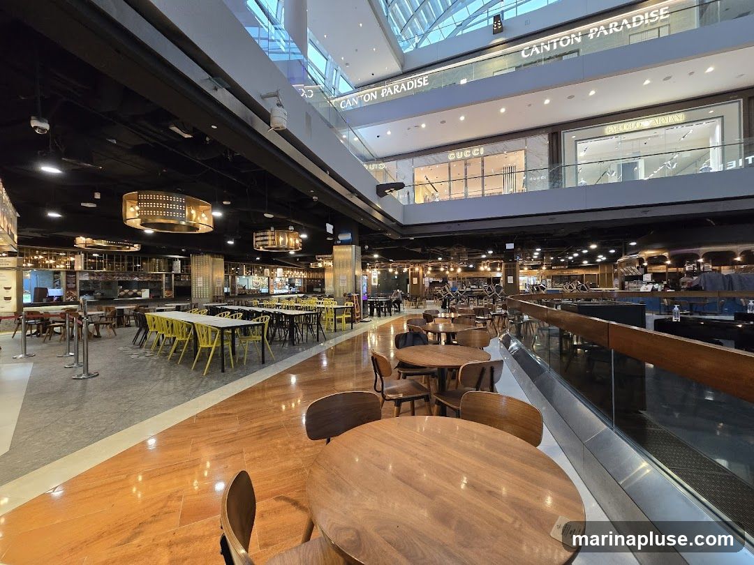 A wide shot of a modern food court with various seating areas and shops visible on different levels. The foreground features round wooden tables and chairs. In the mid-ground, there are long tables with yellow and black chairs, and food stalls with signage. The background shows shops like Gucci and Emporio Armani on an upper level, with a sign for Canton Paradise visible. The overall ambiance is bright and spacious, with a mix of polished wooden floors and tiled areas.