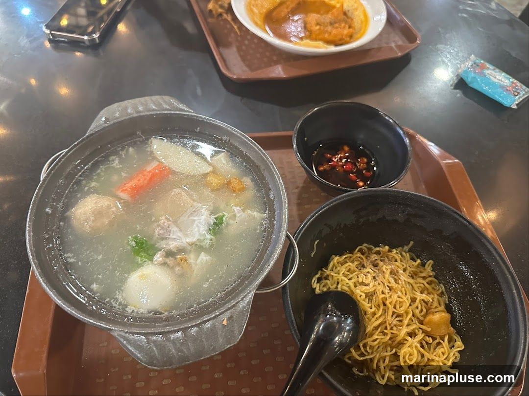 A close-up overhead shot of a meal on a dark table. In the foreground, a black bowl contains a portion of dry noodles with some seasoning. To the left, a metal pot holds a clear soup with various ingredients including meatballs, crab stick, and vegetables. A small black bowl with a dark dipping sauce containing chili flakes sits between the noodle bowl and the soup pot. In the background, another tray holds a dish with curry and a piece of bread or pastry. A smartphone is visible in the upper left corner.