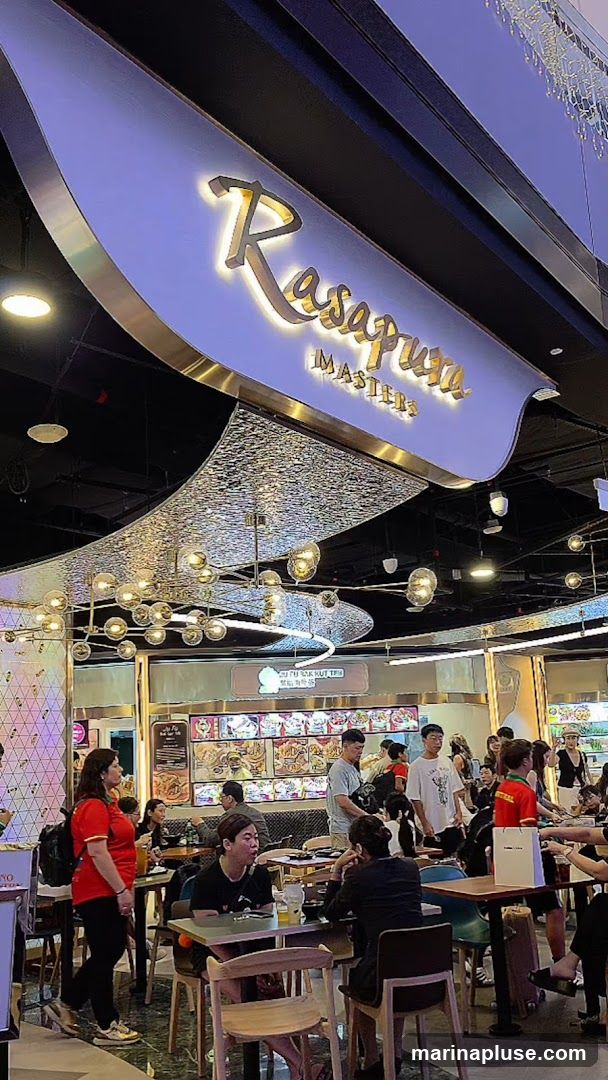 A busy food court with many people dining and queuing. A prominent sign above reads "Rasapura MASTERS". The ceiling features decorative lighting.