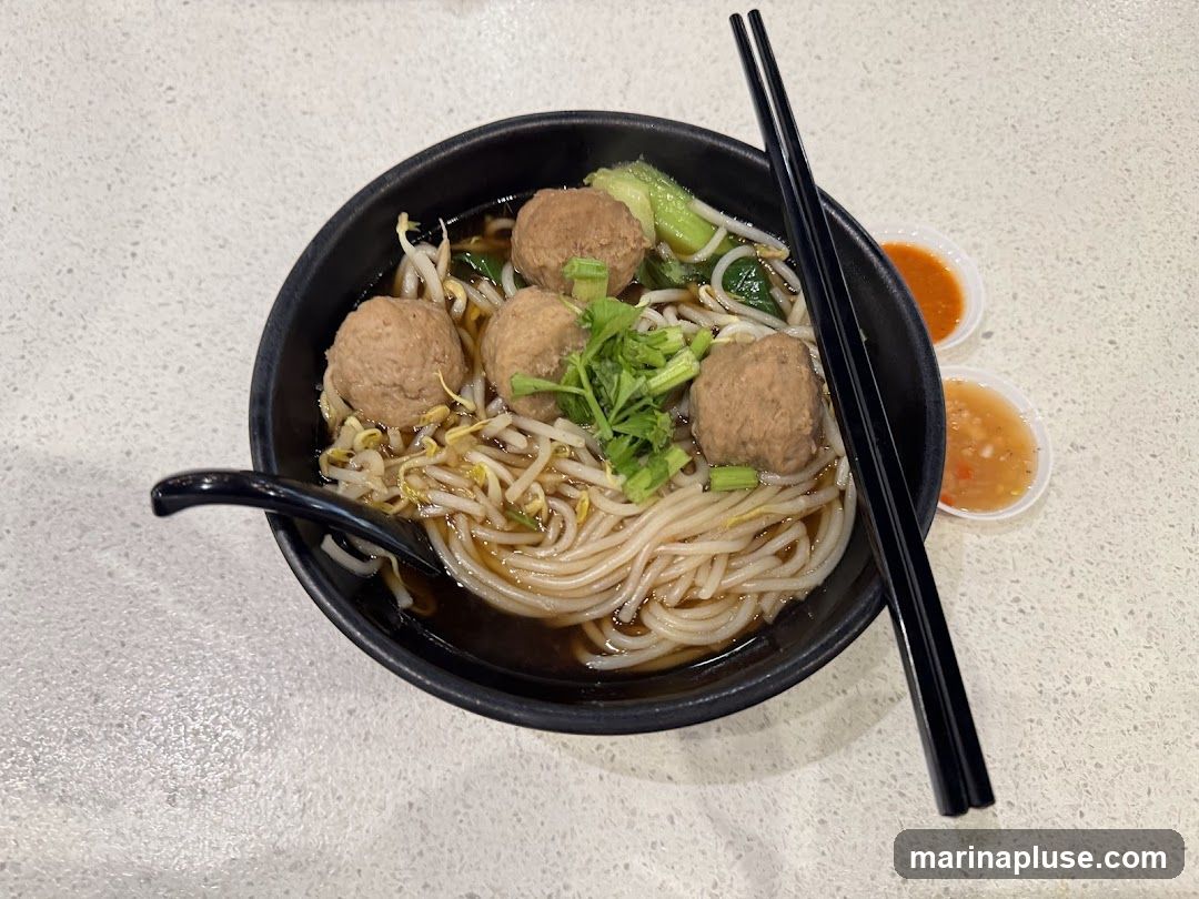 A bowl of noodle soup with meatballs, bean sprouts, and greens, served with two small dipping sauces and chopsticks.