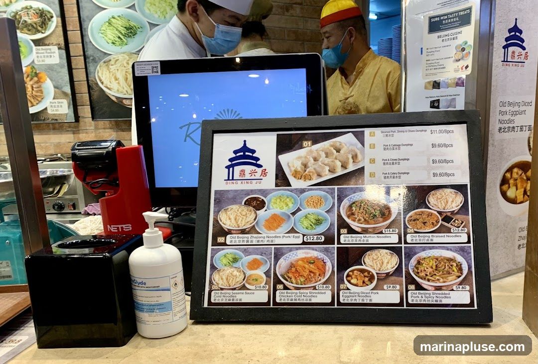 A close-up view of a menu board displaying various Beijing cuisine dishes, including dumplings and noodles. In the background, two chefs are visible, one wearing a white uniform and mask, and the other in a yellow uniform and hat, also wearing a mask. A computer monitor and a hand sanitizer dispenser are also present.