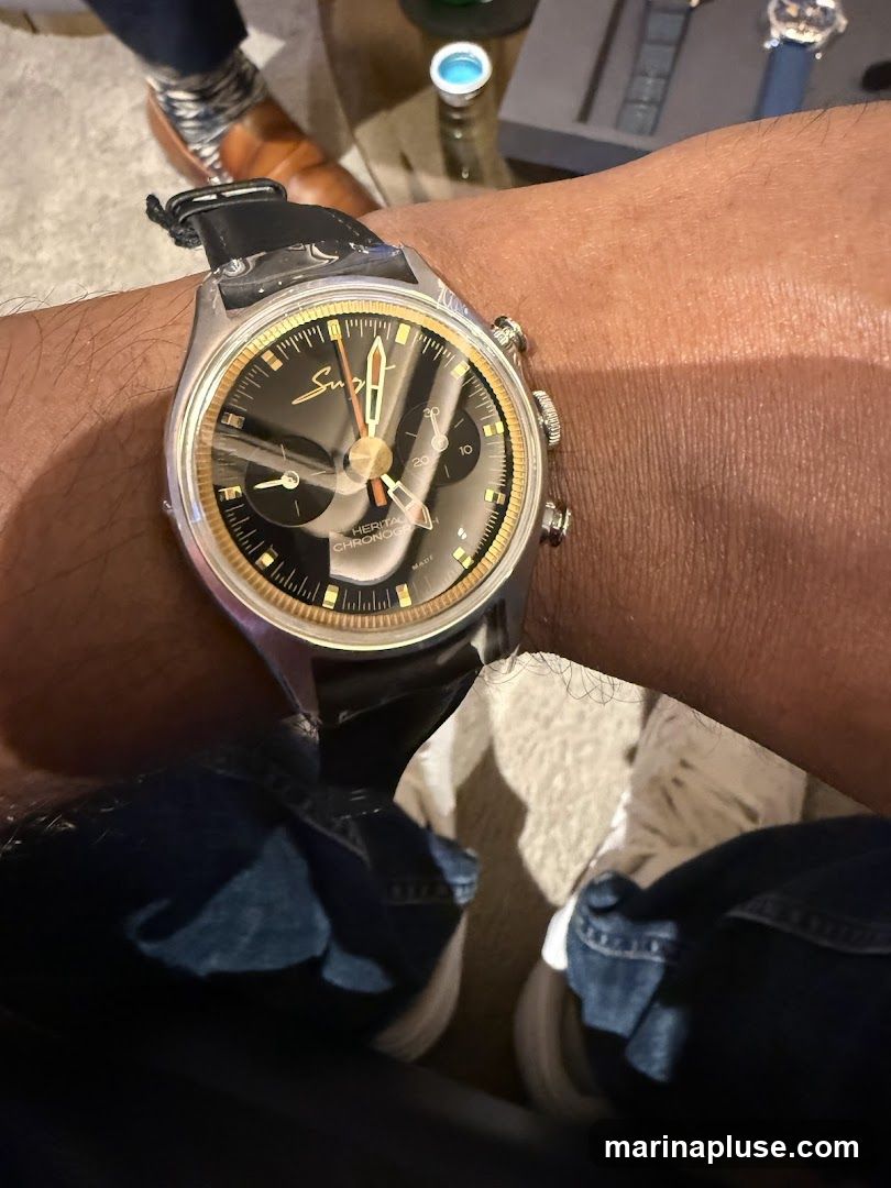 A close-up shot of a person's wrist wearing a vintage-style chronograph watch. The watch has a black dial with gold accents, including the hour markers and hands. The brand name 'Suga' is visible on the dial, along with 'HERITAGE CHRONOGRAPH'. The watch is on a black leather strap. The background shows parts of the person's legs in jeans and shoes, and a blurred background with what appears to be a table and other objects.