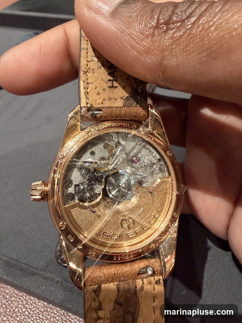 A close-up shot of the intricate mechanical movement of a rose gold watch, with a cork-style strap. The watch case back is transparent, revealing the gears and components. The brand name 'GRONEFELD' is visible on the movement plate. Protective plastic film is still on the watch.