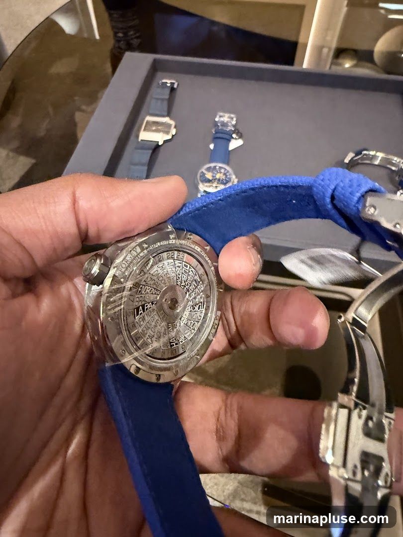 A person's hand holds a watch with a blue strap. The watch face displays a world time complication with various city names. In the background, other watches are displayed on a gray tray.