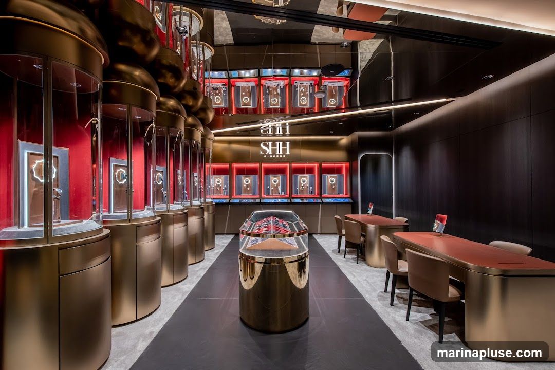 The interior of a high-end watch store features rows of cylindrical display cases with watches, a central display case, and seating areas with tables. The store has a modern and luxurious design with gold accents, red backdrops for the displays, and dark wood paneling on the walls. The ceiling is reflective, adding to the opulent feel. The brand logo "SHH SINCERE" is visible on the wall behind the central display.