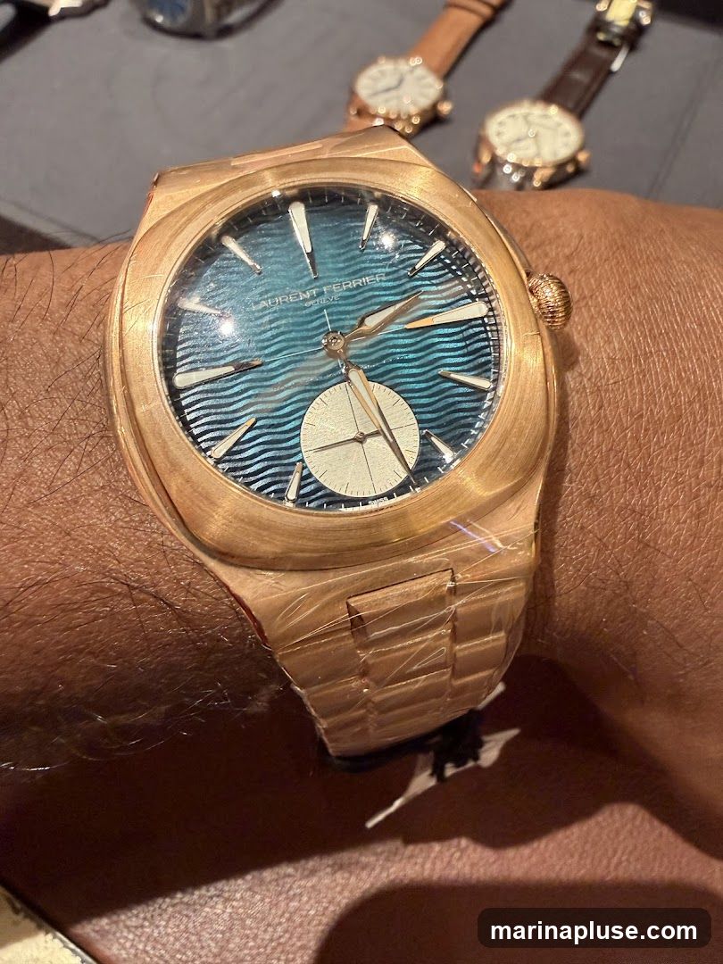 A close-up shot of a rose gold watch with a distinctive blue wave-patterned dial. The watch is on a person's wrist, and the bracelet is still partially covered in protective plastic. The dial features applied hour markers and a small seconds subdial at the 6 o'clock position. The brand name 'LAURENT FERRIER GENEVE' is visible on the dial. In the background, two other watches are out of focus.