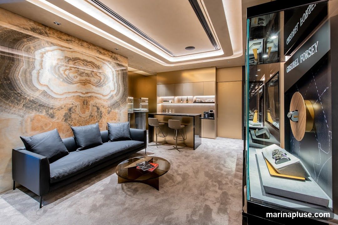 A sophisticated interior space with a large, abstract patterned marble wall, a dark grey sofa with cushions, and a glass coffee table. In the background, there is a bar area with stools and a coffee machine. To the right, a display case showcases luxury watches, with the brand name "GREUBEL FORSEY" visible.