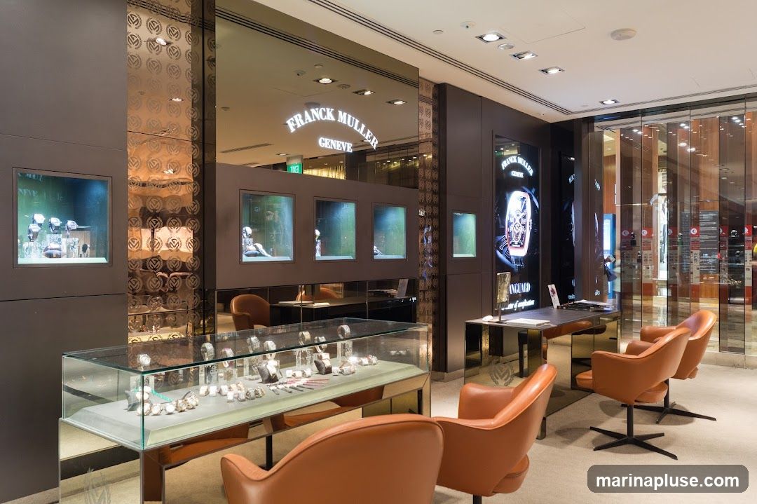The interior of a Franck Muller watch store, featuring display cases filled with watches, illuminated display windows showcasing watches and mannequins, and comfortable seating areas with orange leather chairs. A large digital display shows a Franck Muller watch. The store has a luxurious and modern design with mirrored surfaces and warm lighting.