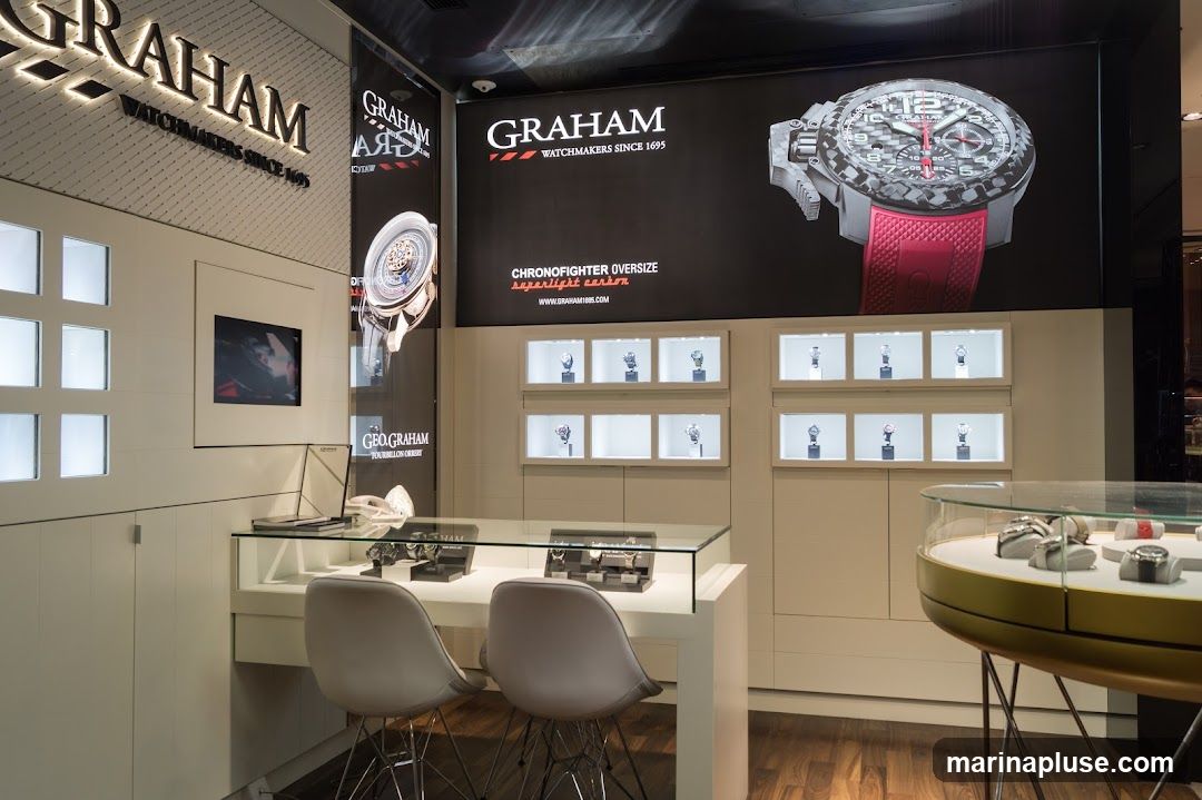 The interior of a Graham watch store, featuring a large display screen showing a Graham Chronofighter Oversize watch. The store has illuminated display cases showcasing various watches, a glass counter with watches on display, and two chairs. The Graham logo and "Watchmakers Since 1695" are prominently displayed on the wall and signage.