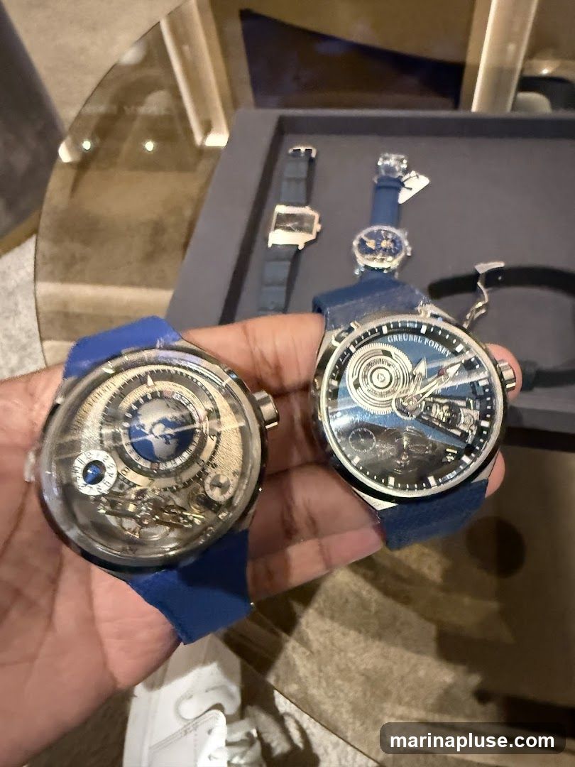A person is holding two luxury watches with blue straps. The watches have intricate mechanical movements visible through their faces. In the background, other watches are displayed in a tray on a glass table.