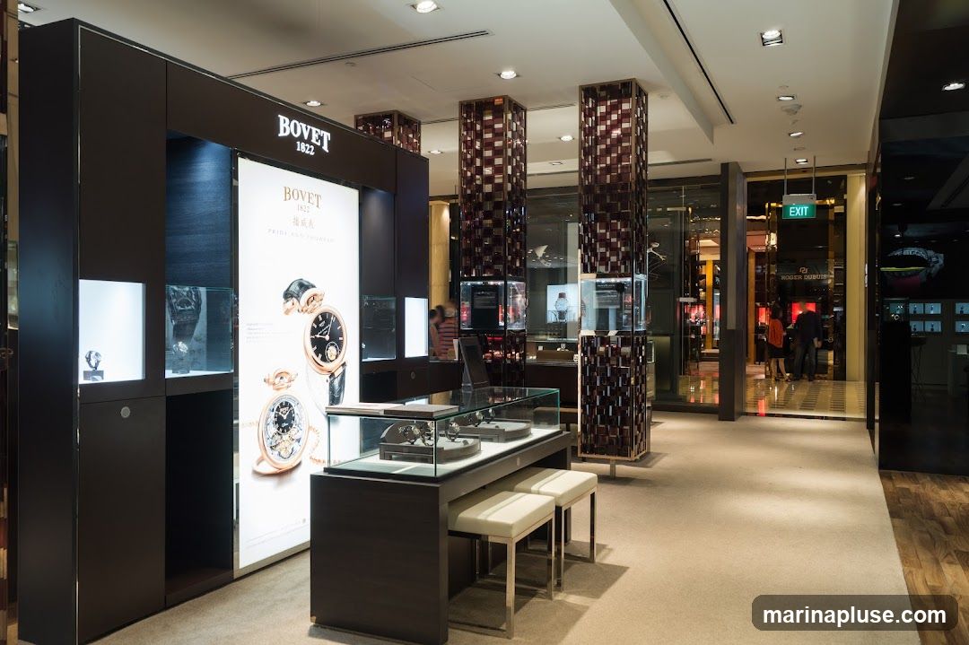 A well-lit interior of a luxury watch store, featuring a prominent display for Bovet watches. The display includes illuminated showcases with watches and a large backlit advertisement showcasing two Bovet timepieces. In the foreground, a glass display case with watches sits on a dark wood counter, accompanied by two white stools. Further into the store, decorative pillars and other watch displays are visible, with an exit sign above a doorway leading to another section of the store.