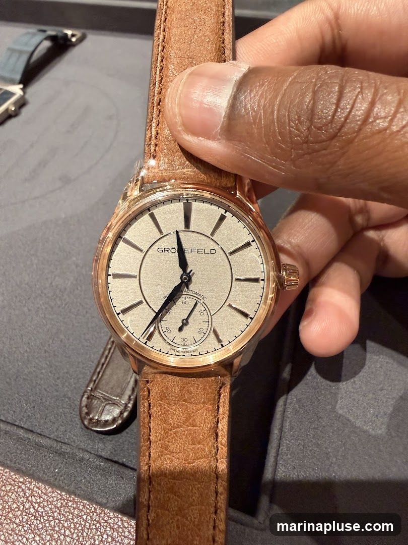 A close-up shot of a Gronfeld watch with a light-colored dial and a brown leather strap. The watch has a rose gold case and a small seconds subdial. The brand name "GRÖNEFELD" is visible on the dial. A person's hand is holding the watch. Another watch strap is visible in the background.
