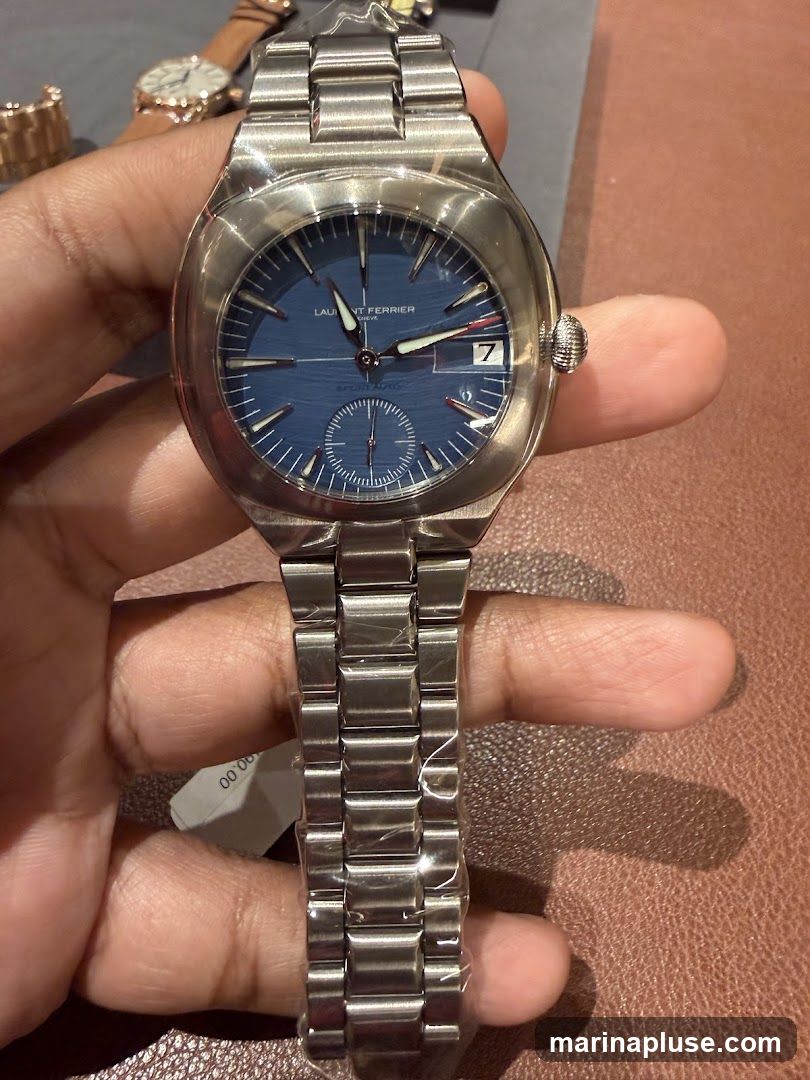 A person is holding a stainless steel watch with a blue dial. The watch has a metal bracelet and a date window. The brand name "LAURENT FERRIER GENEVE" is visible on the dial. There are other watches in the background.