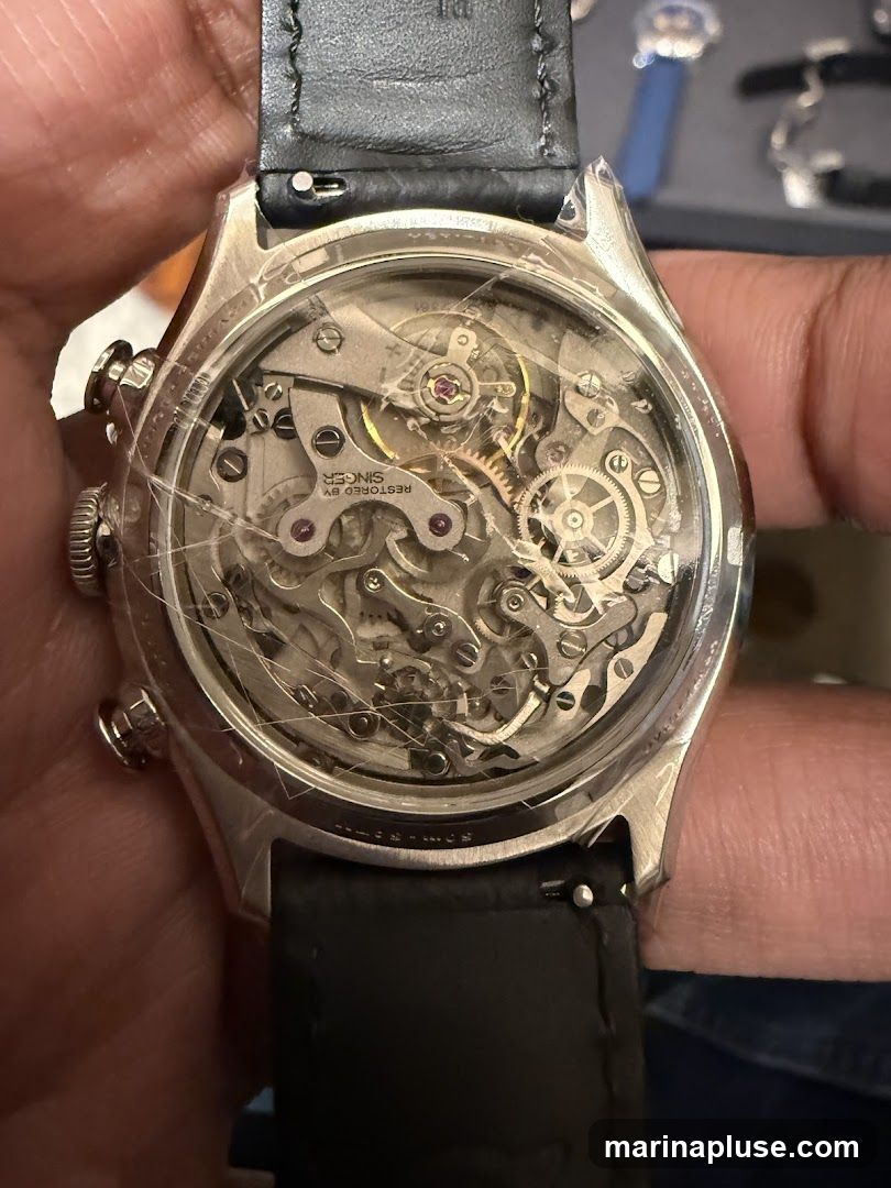 A close-up view of the intricate mechanical movement of a watch, seen through a transparent case back. The movement features numerous gears, springs, and jewels, with text visible on some components, including "RESTORED BY SINGER". The watch has a black leather strap.