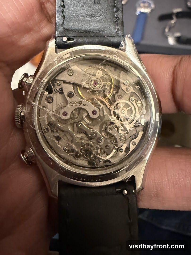 A close-up view of the intricate mechanical movement of a watch, seen through a transparent case back. The movement features numerous gears, springs, and jewels, with text visible on some components, including "RESTORED BY SINGER". The watch has a black leather strap.