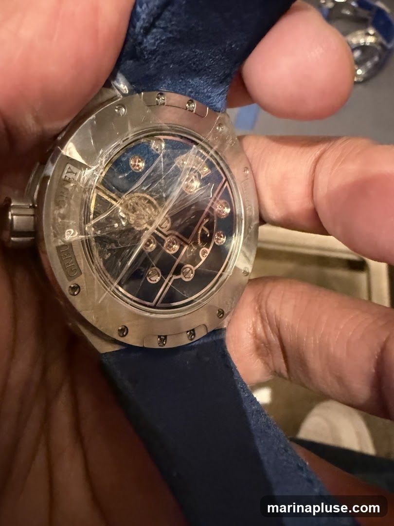 A close-up shot of the back of a watch, held by a person's fingers. The watch has a silver-toned case with visible screws around the edge. The caseback is transparent, revealing the intricate inner workings of the watch movement. The dial appears to be dark blue with some metallic accents. A blue strap is attached to the watch. The protective plastic film is still on the caseback, showing some scratches.
