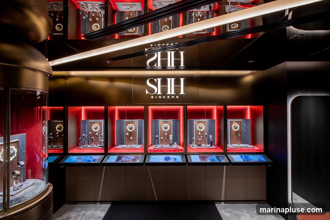 A well-lit interior display of luxury watches in a modern retail setting. The display features multiple illuminated showcases, each presenting several watches on stands. Below the showcases are digital screens displaying watch imagery. The brand name "SHH SINCERE" is prominently displayed above the watch displays. The overall aesthetic is sleek and sophisticated, with a color palette of gold, black, and red accents.