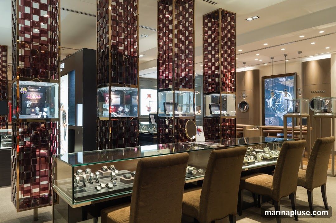 The interior of a high-end watch store, featuring illuminated display cases filled with watches. Tall, decorative pillars with a checkered pattern of red and gold accents line the space. In the background, a large digital screen displays a close-up of a watch face, with the brand name "PANERAI" visible. Seating areas with plush chairs are arranged around display counters.