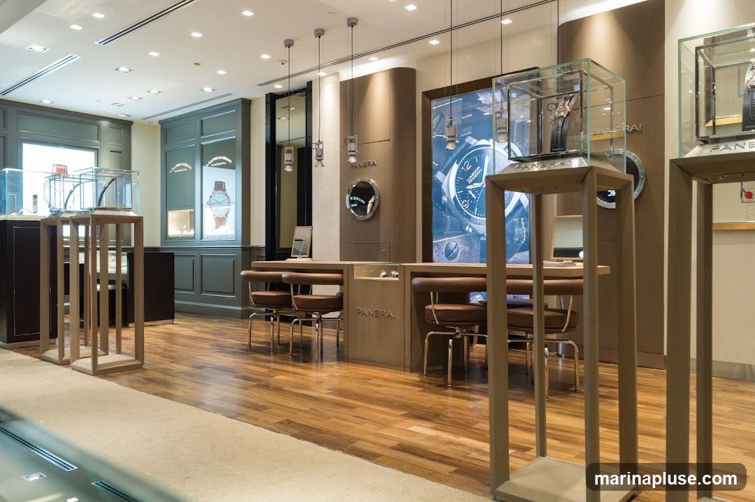 The interior of a high-end watch store, featuring display cases with watches, a central counter with seating, and large promotional imagery on the walls. The store has polished wooden floors and elegant lighting.