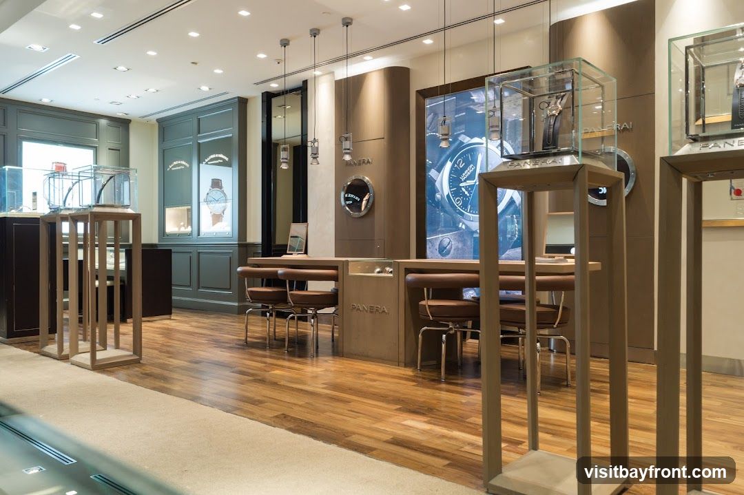 The interior of a high-end watch store, featuring display cases with watches, a central counter with seating, and large promotional imagery on the walls. The store has polished wooden floors and elegant lighting.