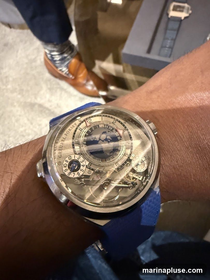 A close-up shot focuses on a complex luxury watch with a blue strap worn on a person's wrist. The watch face displays multiple dials and intricate mechanical components, with the brand name "GREUBEL FORSEY" visible. In the background, a person's legs are visible, wearing brown loafers and patterned socks, suggesting a formal or semi-formal setting.