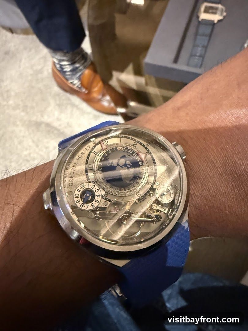 A close-up shot focuses on a complex luxury watch with a blue strap worn on a person's wrist. The watch face displays multiple dials and intricate mechanical components, with the brand name "GREUBEL FORSEY" visible. In the background, a person's legs are visible, wearing brown loafers and patterned socks, suggesting a formal or semi-formal setting.