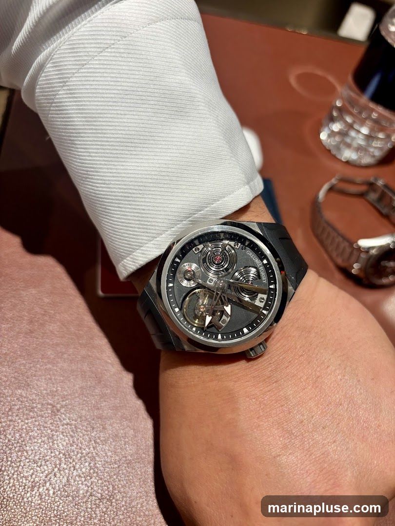A close-up shot focuses on a man's wrist, adorned with a sophisticated mechanical watch. The watch face displays intricate gears and mechanisms, suggesting a high-end timepiece. The man is wearing a white dress shirt with textured sleeves. In the background, a bottle of water and another watch are visible on a brown surface.