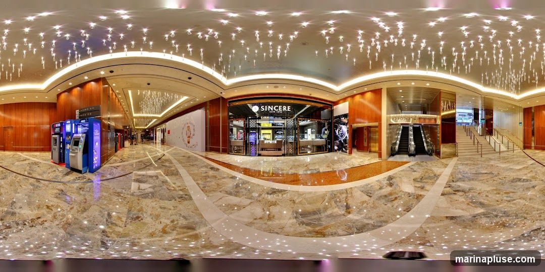 A panoramic view of a luxurious shopping mall interior. The floor is made of polished marble with intricate patterns. The ceiling is adorned with numerous sparkling lights, creating a dazzling effect. On the left, there are several ATMs and information kiosks. In the center, a prominent storefront with the name "SINCERE ESTD 1954" is visible, displaying watches and jewelry. To the right, escalators and a grand staircase lead to upper levels. The overall ambiance is upscale and modern.