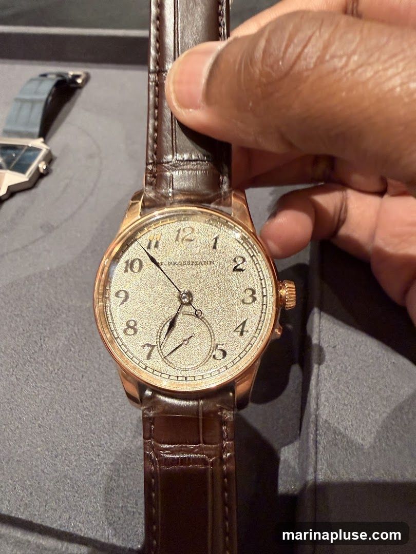 A person's hand is holding a rose gold watch with a textured champagne dial and Arabic numerals. The watch has a brown alligator leather strap. In the background, another watch with a geometric design and a blue strap is visible. The watches are displayed on a gray surface.