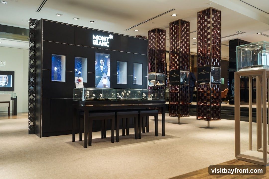 The interior of a Montblanc store, featuring display cases filled with watches and other luxury items. The prominent Montblanc logo is visible above a display wall. Several tall, decorative pillars with glass shelves are also present, showcasing more merchandise. The store has a clean, modern aesthetic with carpeted floors and warm lighting.