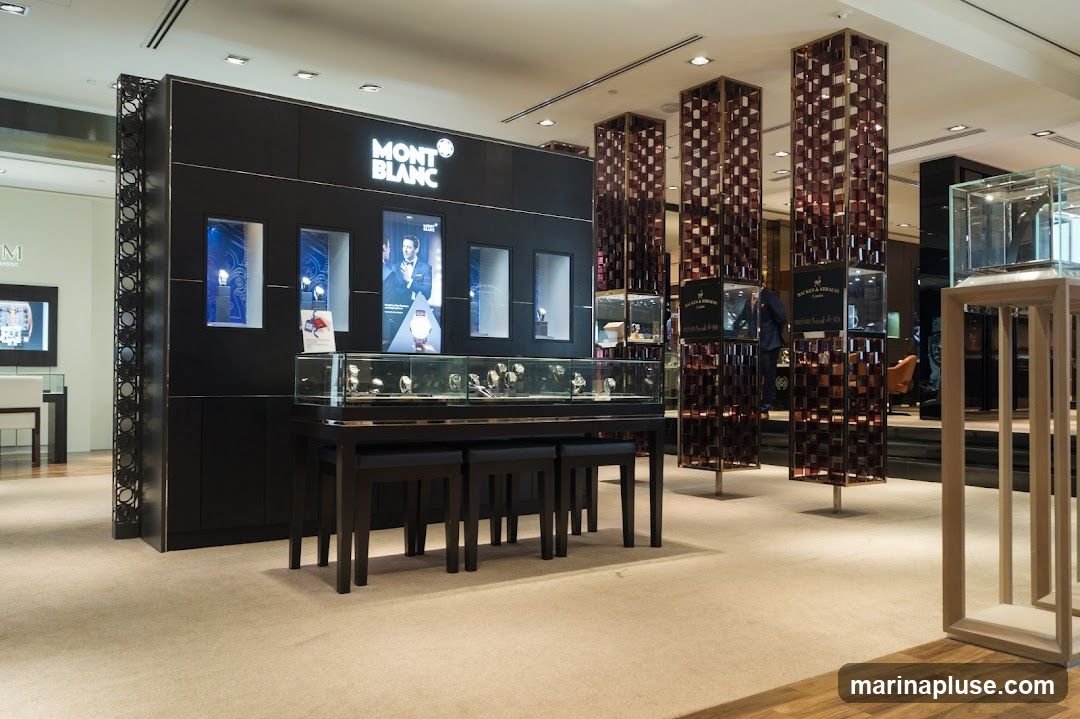 The interior of a Montblanc store, featuring display cases filled with watches and other luxury items. The prominent Montblanc logo is visible above a display wall. Several tall, decorative pillars with glass shelves are also present, showcasing more merchandise. The store has a clean, modern aesthetic with carpeted floors and warm lighting.