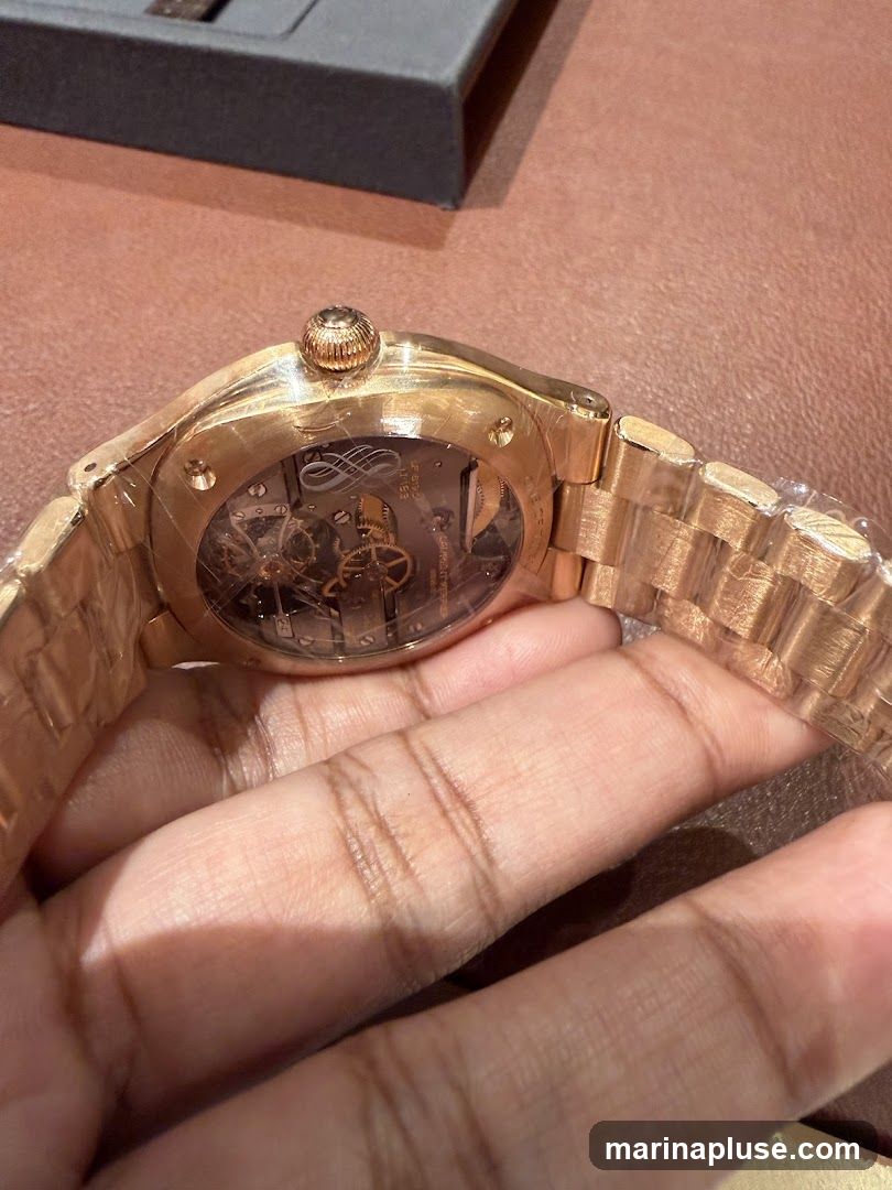 A close-up shot of a rose gold watch with a skeleton back, revealing the intricate inner workings of the timepiece. The watch is held by a hand, and a dark gray box is visible in the background. The watch appears to be new, with protective plastic still on the band and case.