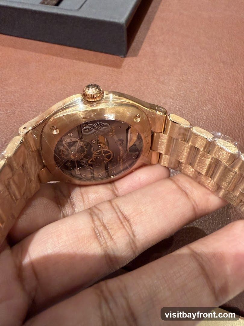 A close-up shot of a rose gold watch with a skeleton back, revealing the intricate inner workings of the timepiece. The watch is held by a hand, and a dark gray box is visible in the background. The watch appears to be new, with protective plastic still on the band and case.