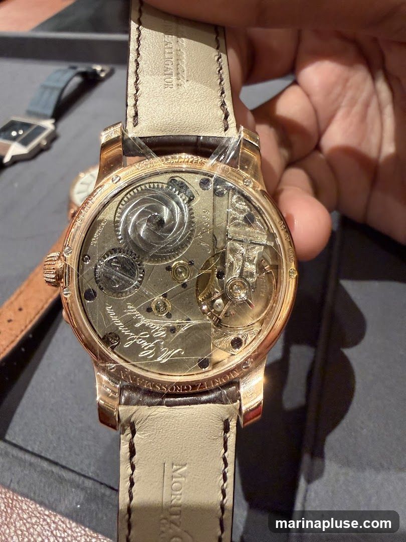 A close-up view of the intricate mechanical movement of a rose gold watch. The watch has a light-colored leather strap with visible stitching and branding. The movement features detailed gears, springs, and engraved text. Another watch with a blue strap and a geometric face is visible in the blurred background, along with a portion of another watch with a white face.