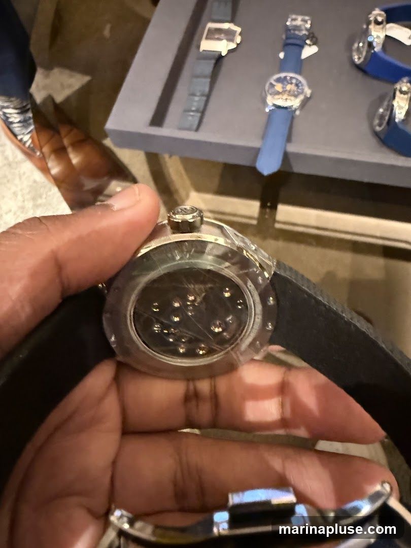 A person's hands are holding a watch, showing the intricate mechanical movement through a transparent case back. In the background, several other watches are displayed on a grey tray, including one with a blue strap and a rectangular face, and another with a round face and a blue strap. A pair of brown dress shoes and dark trousers are visible in the lower left corner.