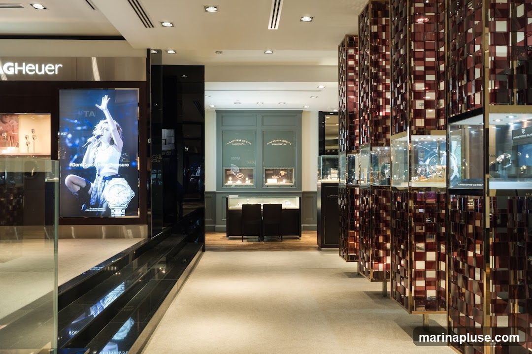 The interior of a high-end watch store. A large digital display features a woman and a watch with the text "#DontCrackUnderPressure". To the right, a decorative screen with reflective panels displays watches in glass cases. In the background, a display cabinet with two chairs in front of it shows more watches, with signage for "J. LAPORTE & CIE".