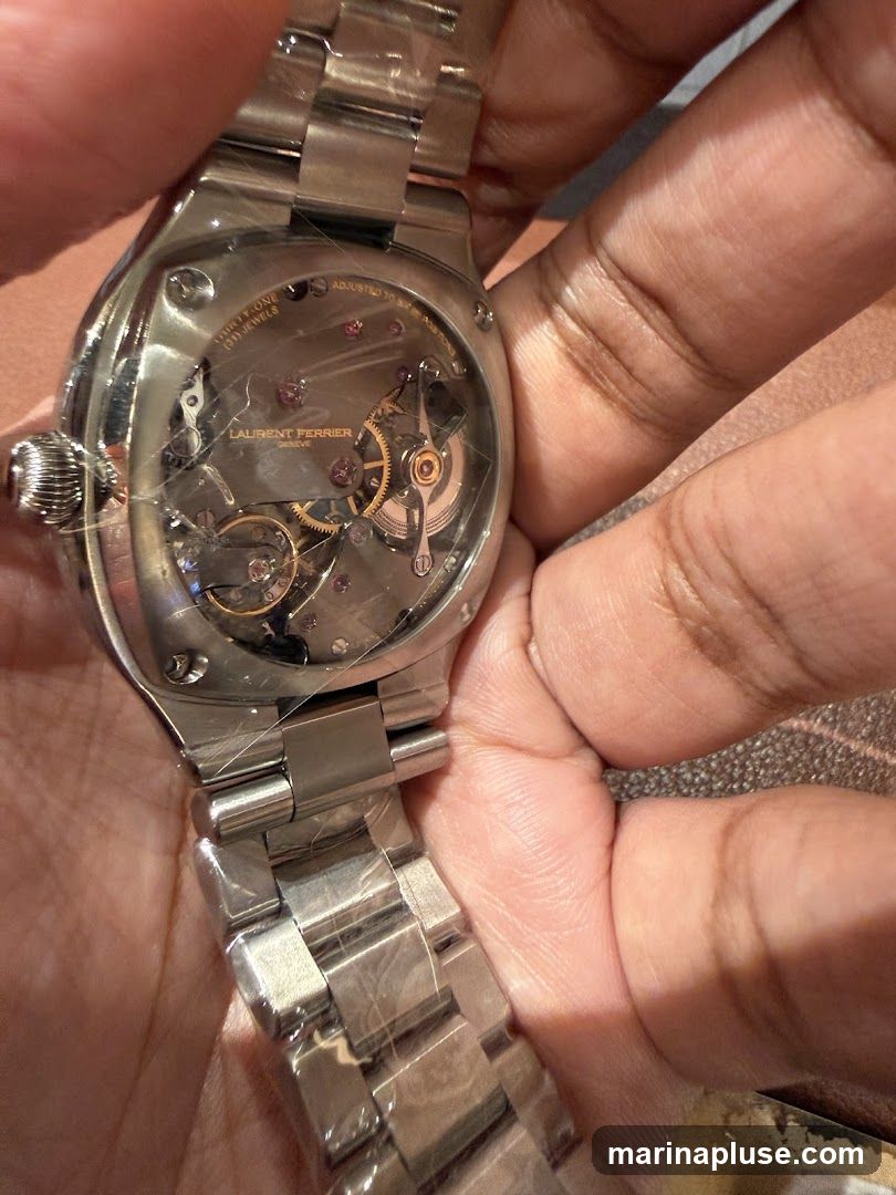 A close-up view of the back of a watch, showcasing its intricate mechanical movement. The watch has a stainless steel band and case. The brand name "LAURENT FERRIER GENEVE" is visible on the movement. The watch appears to be new, with protective plastic still on the band and case.