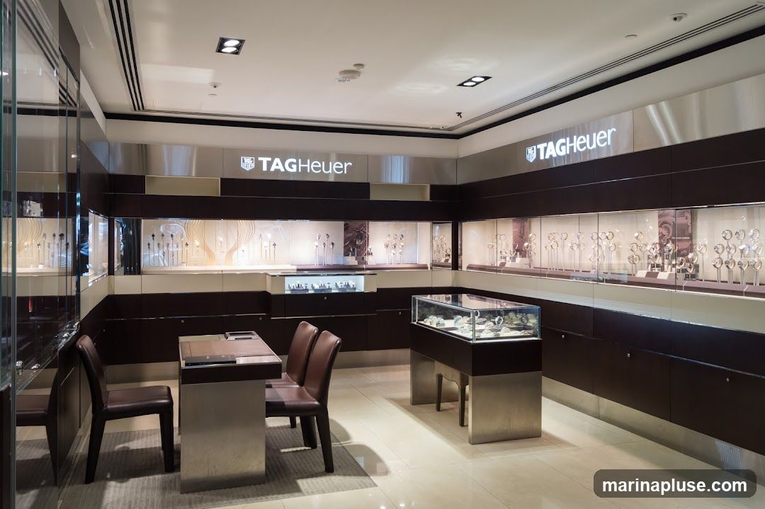 The interior of a TAG Heuer watch store, featuring illuminated display cases filled with watches. The store has a modern and luxurious design with dark wood accents and polished metal finishes. There are seating areas with chairs and a table for customers. The TAG Heuer logo is prominently displayed on the walls.