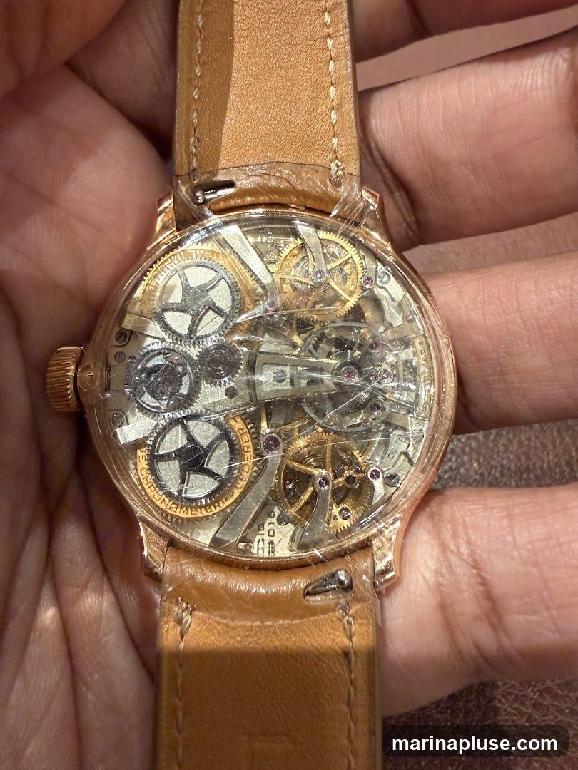 A close-up shot of a skeletonized watch with a rose gold case and a tan leather strap. The watch face reveals intricate mechanical gears and components, with some text visible on the inner bezel. The watch appears to be new, with protective plastic still on the crystal.
