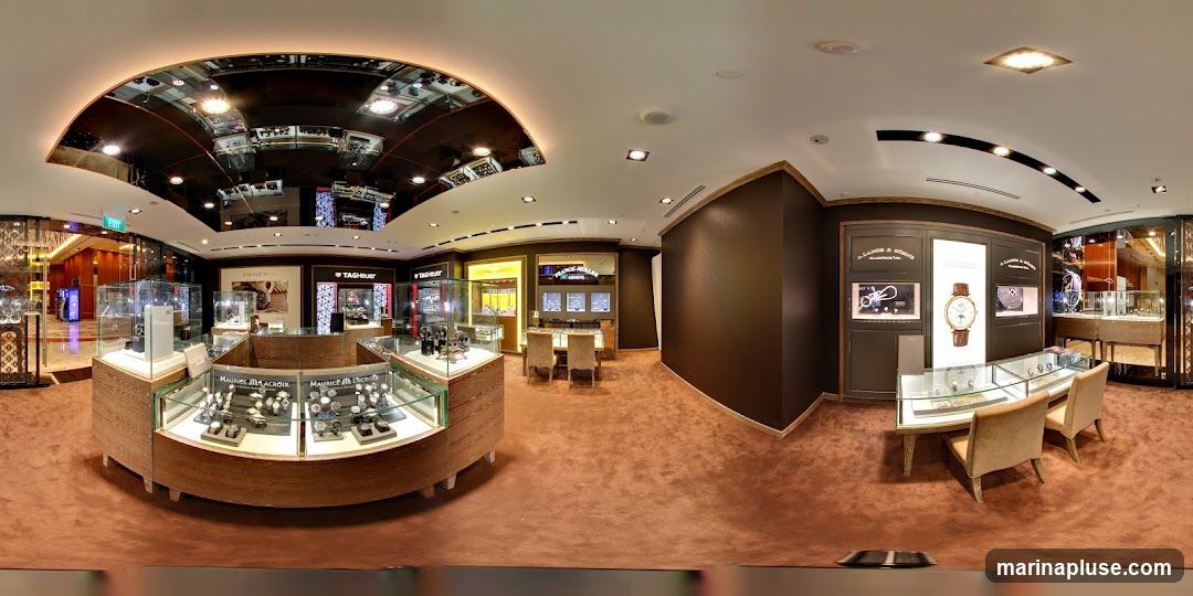 A panoramic view of the interior of a luxury watch store. Display cases filled with watches are arranged throughout the space. Brands such as TAG Heuer and Maurice Lacroix are visible on signage and displays. The store features plush carpeting, elegant lighting, and seating areas for customers.