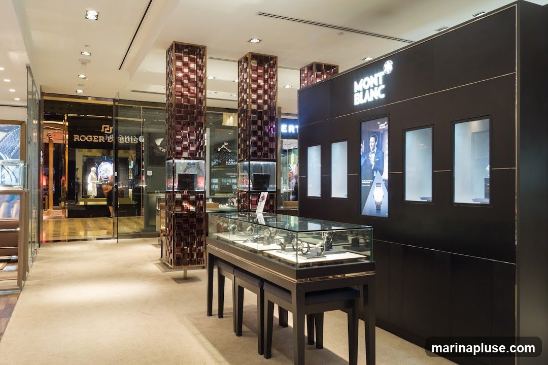 The interior of a luxury watch store, featuring a prominent Montblanc display wall with illuminated windows and a large advertisement. In the foreground, a glass display case showcases various watches. To the left, another store with a "ROGER DUBUIS" sign is visible through a glass facade. The store has a plush carpeted floor and decorative pillars.