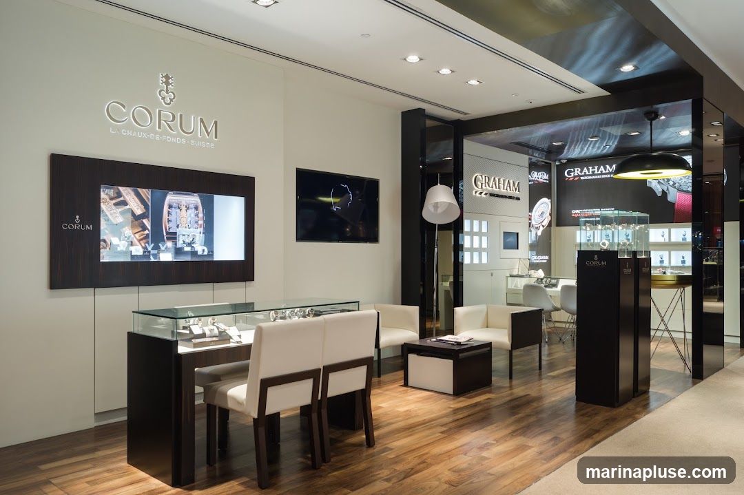 The interior of a luxury watch store featuring displays for Corum and Graham watches. The store has a modern and elegant design with wooden floors, white seating areas, and glass display cases showcasing watches. A large screen displays Corum advertisements.