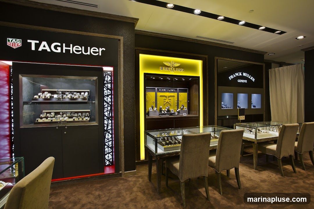 A well-lit interior of a luxury watch store, featuring displays for TAG Heuer, Breitling, and Franck Muller. Several glass display cases showcase a variety of watches, with plush seating areas for customers. The overall ambiance is sophisticated and upscale.