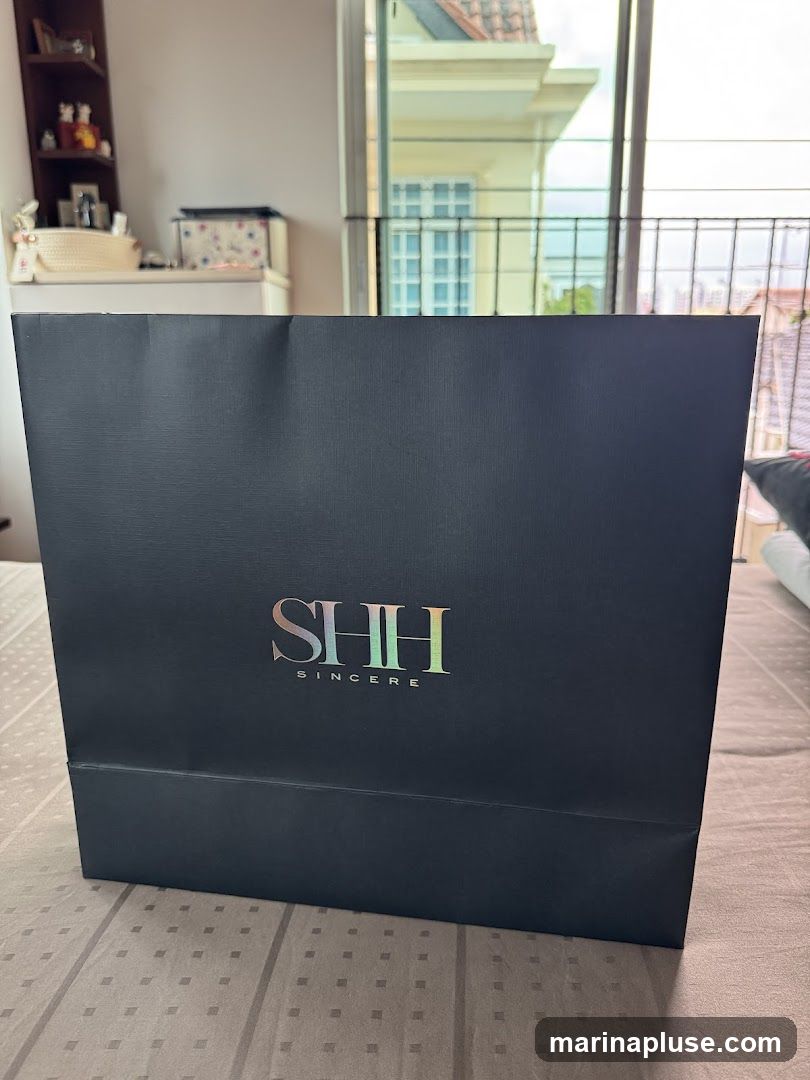 A black paper shopping bag with the iridescent logo "SHH SINCERE" printed on it. The bag is placed on a bed or sofa. In the background, a window shows a balcony and buildings outside.
