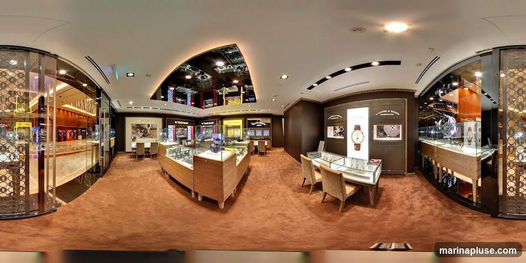 A panoramic view of the interior of a high-end watch store. Display cases filled with watches are arranged throughout the space. The store features plush brown carpeting, elegant lighting, and mirrored ceilings. In the background, a walkway or mall area is visible through glass doors. Prominent signage for brands like TAG Heuer and A. Lange & Söhne are displayed.